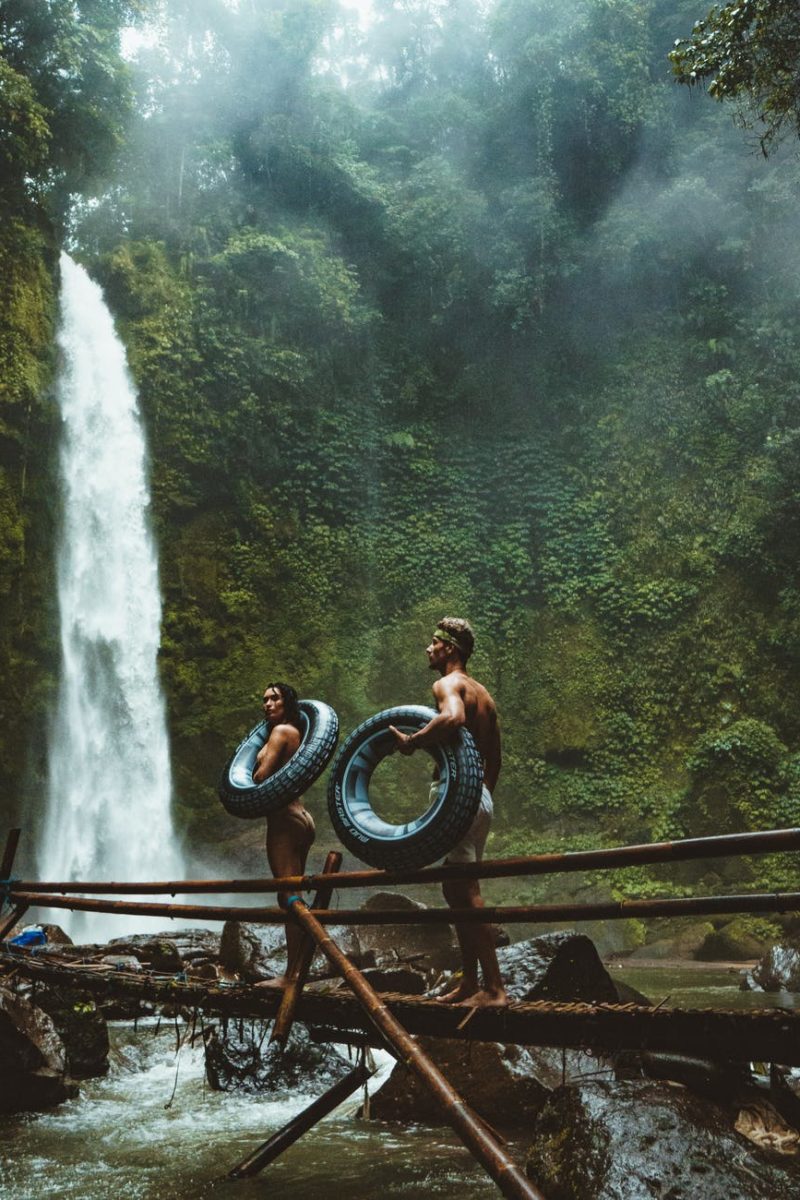 two person carrying black inflatable pool float on brown wooden bridge near waterfalls