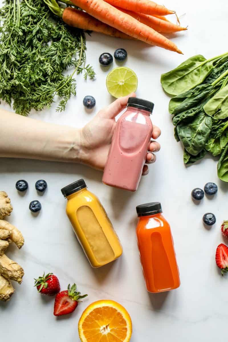 person holding bottle of smoothie beside green leaves carrots and berries