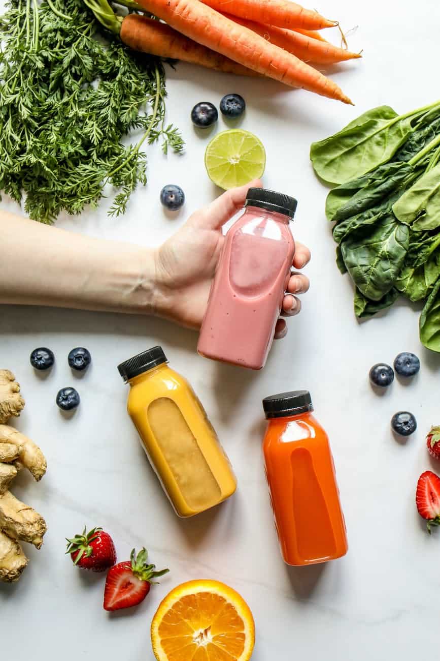 person holding bottle of smoothie beside green leaves carrots and berries