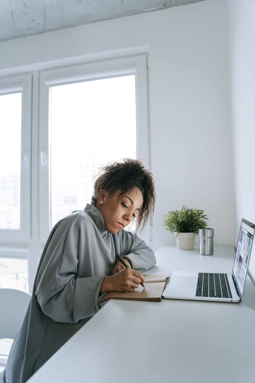 a woman writing on her notebook in front of her laptop