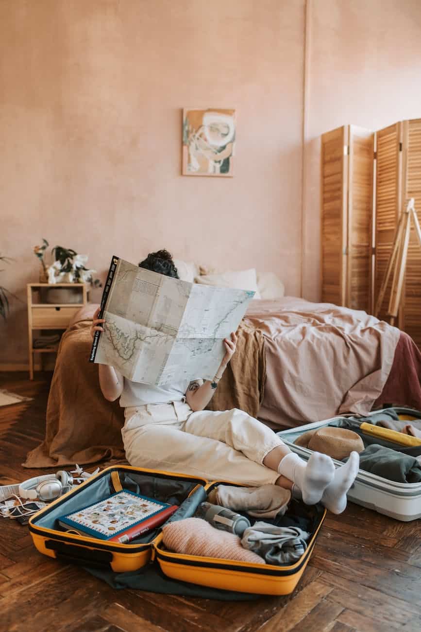 person in white pants lying on bed with a suitcase in front of them and a map in their hands