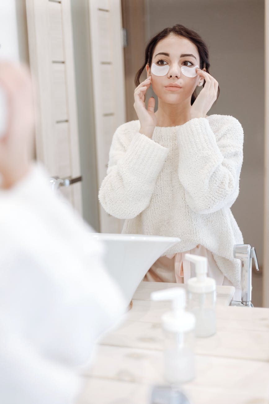 woman in white sweater wearing under eye masks touching her face while looking at her own reflection on the mirror