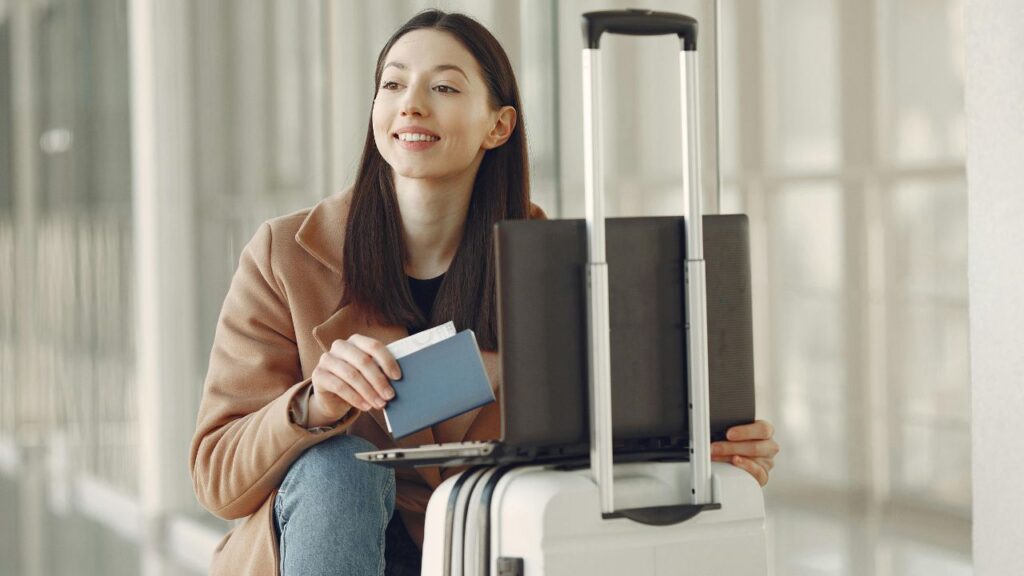 Traveler holding a passport and boarding pass while sitting next to a suitcase and laptop at the airport.