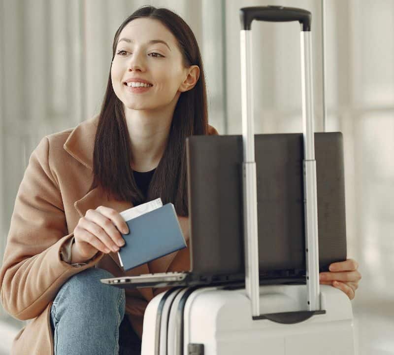 Traveler holding a passport and boarding pass while sitting next to a suitcase and laptop at the airport.