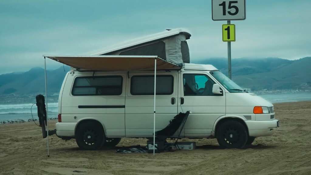 A white camper van with an extended awning parked on a sandy beach, with camping gear set up beside it and misty coastal mountains in the background.
