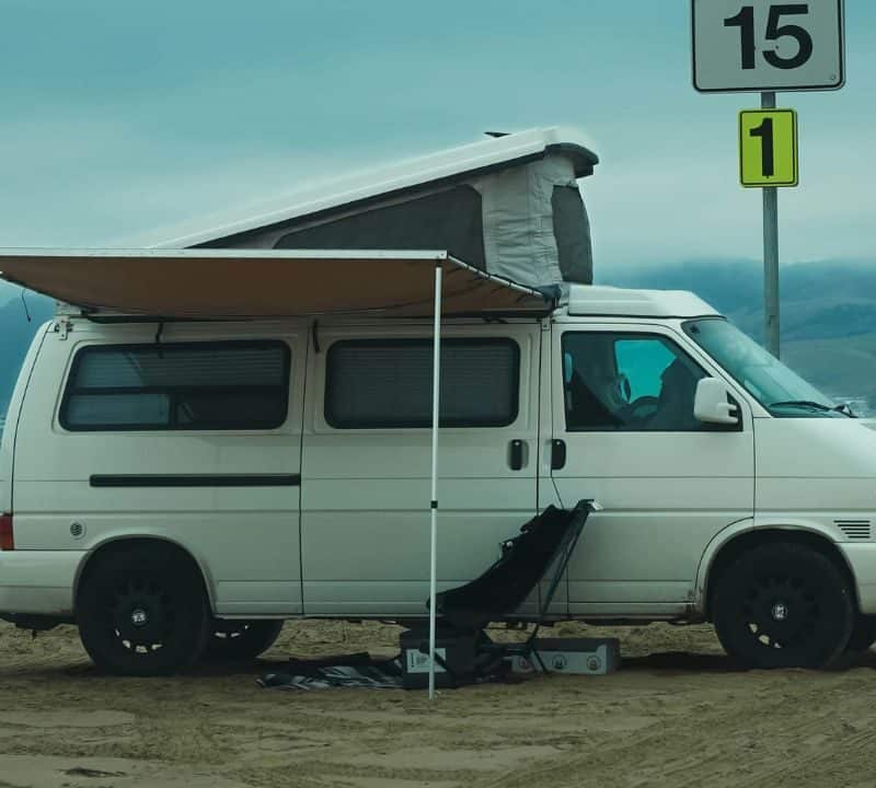A white camper van with an extended awning parked on a sandy beach, with camping gear set up beside it and misty coastal mountains in the background.