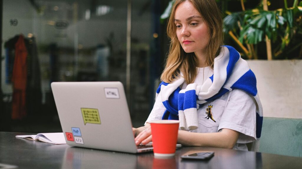 Young woman working remotely on a laptop in a coworking space, wearing a blue and white scarf with a coffee cup nearby.