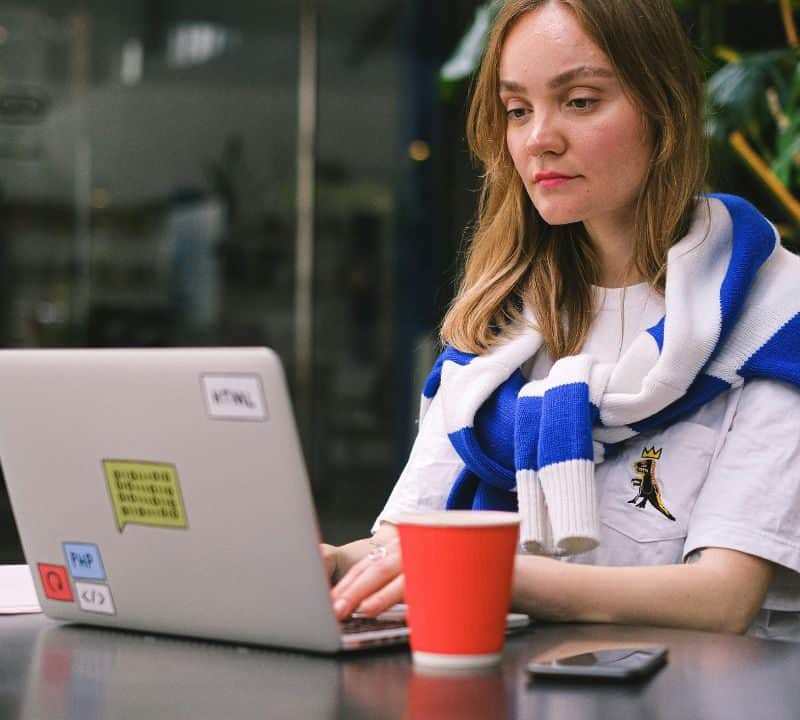 Young woman working remotely on a laptop in a coworking space, wearing a blue and white scarf with a coffee cup nearby.