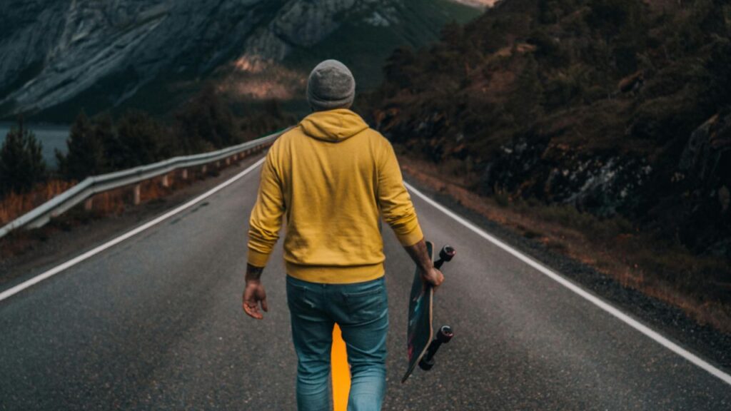 A person in a yellow hoodie and beanie walking alone on a mountain road while holding a skateboard, surrounded by nature and rugged scenery.
