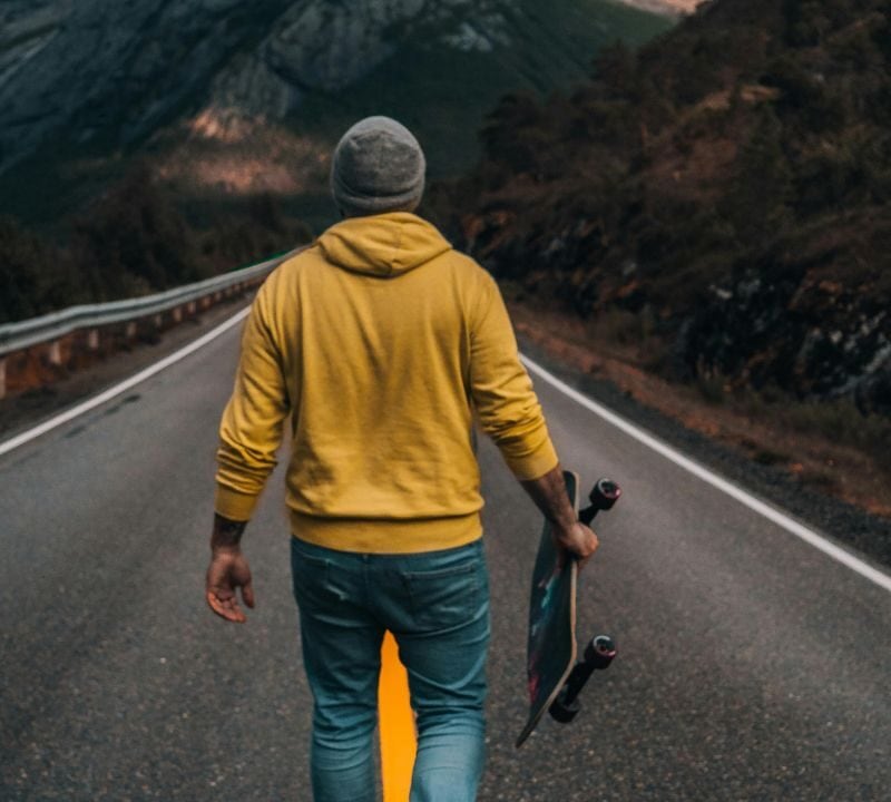 A person in a yellow hoodie and beanie walking alone on a mountain road while holding a skateboard, surrounded by nature and rugged scenery.