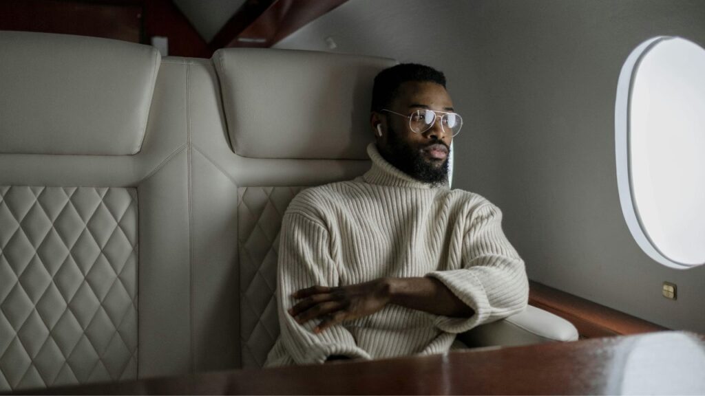 Man sitting comfortably in a premium airplane seat, wearing glasses and earbuds, looking out the window during a long flight.