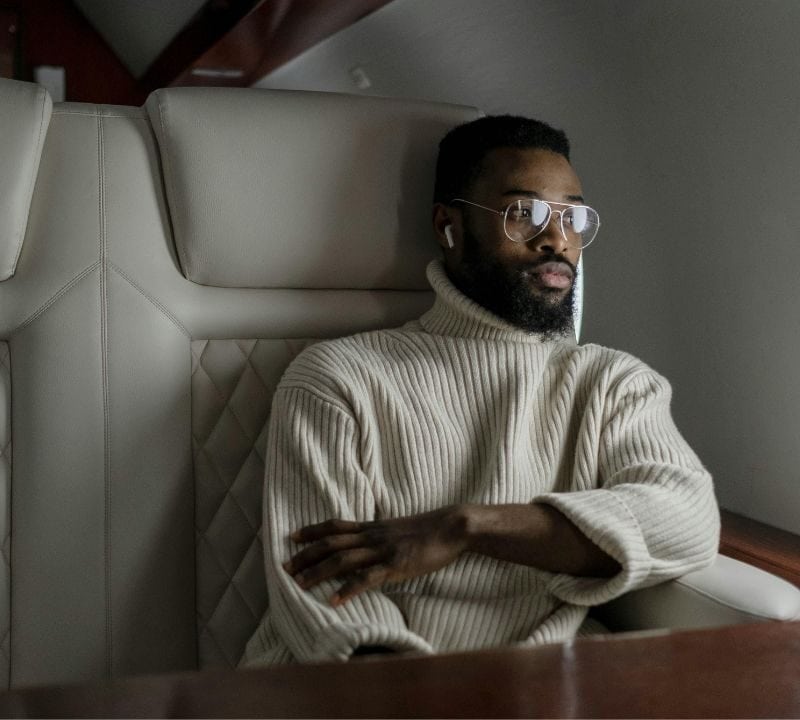 Man sitting comfortably in a premium airplane seat, wearing glasses and earbuds, looking out the window during a long flight.