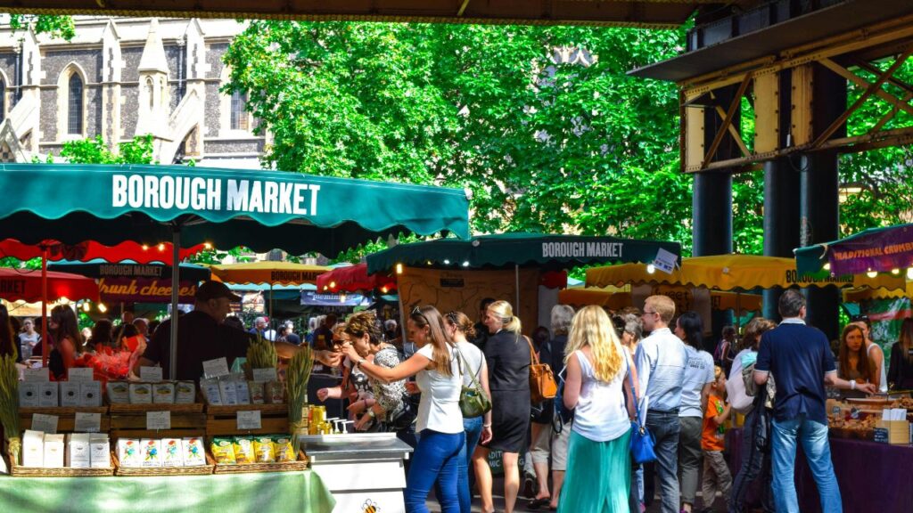 Crowds browsing stalls at Borough Market with green canopies, food vendors, and historic buildings visible in the background on a sunny day.