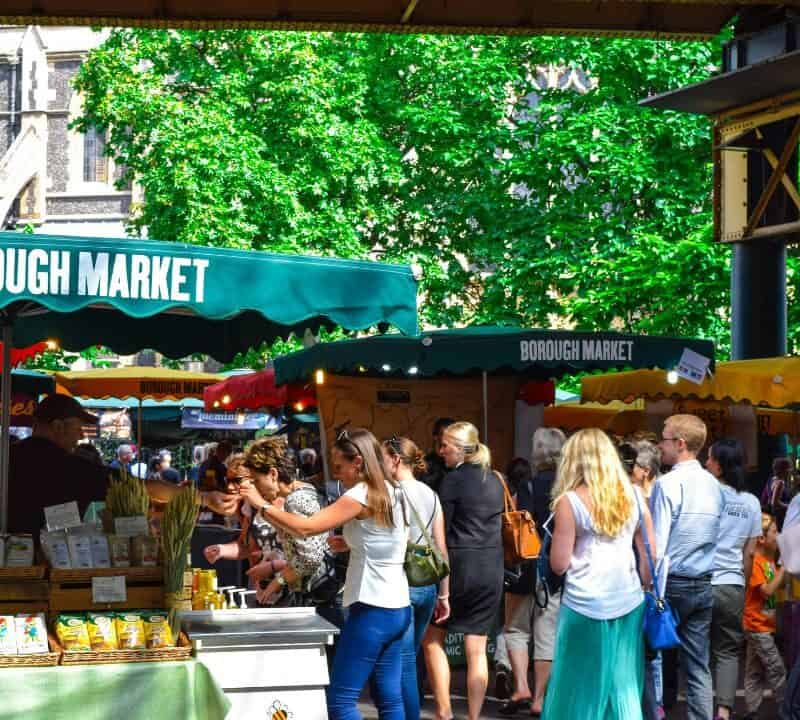 Crowds browsing stalls at Borough Market with green canopies, food vendors, and historic buildings visible in the background on a sunny day.