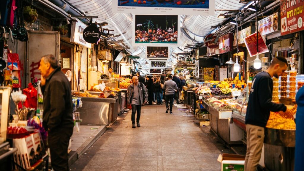 Vibrant scene at Jerusalem’s Mahane Yehuda Market with people walking through a bustling aisle lined with colorful produce, street food stalls, and hanging signs.