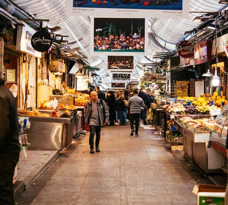 Vibrant scene at Jerusalem’s Mahane Yehuda Market with people walking through a bustling aisle lined with colorful produce, street food stalls, and hanging signs.