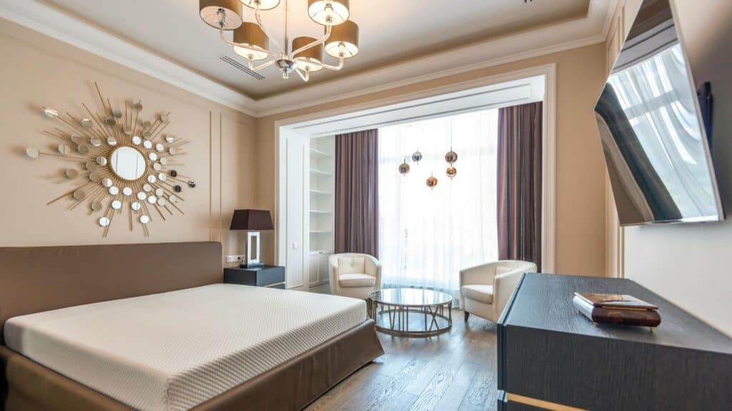 Elegant modern hotel suite with a designer chandelier, sunburst wall mirror, neutral-toned furniture, and large window draped with sheer curtains, creating a bright and sophisticated ambiance.