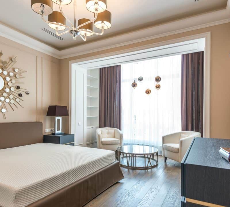 Elegant modern hotel suite with a designer chandelier, sunburst wall mirror, neutral-toned furniture, and large window draped with sheer curtains, creating a bright and sophisticated ambiance.