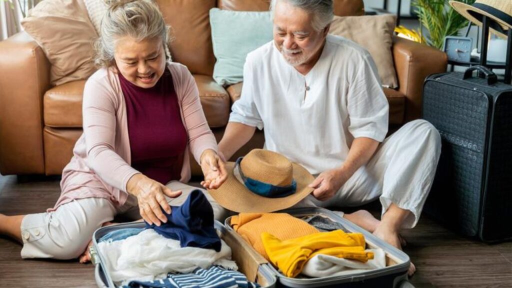 An elderly couple sitting on the floor happily packing clothes and a hat into an open suitcase, with a suitcase and couch in the background.