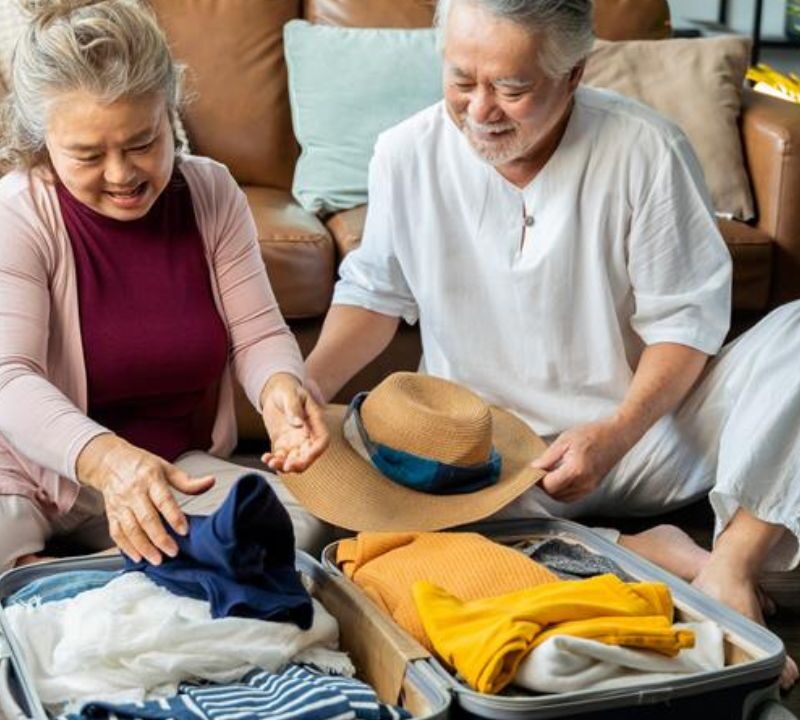 An elderly couple sitting on the floor happily packing clothes and a hat into an open suitcase, with a suitcase and couch in the background.