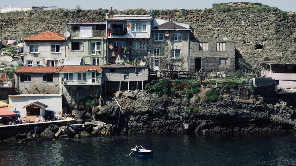 Rustic cliffside homes overlook calm coastal waters with a small rowboat floating below and laundry hanging from balconies.