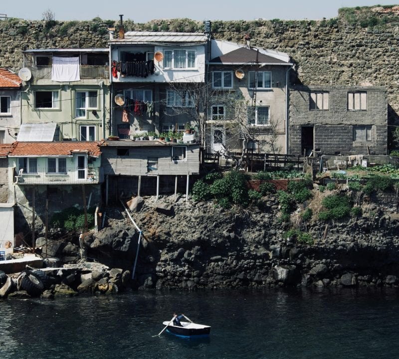 Rustic cliffside homes overlook calm coastal waters with a small rowboat floating below and laundry hanging from balconies.