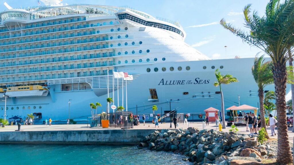 Passengers walk near the massive cruise ship Allure of the Seas docked at a sunny Caribbean port, with palm trees, clear skies, and flags waving near the pier.