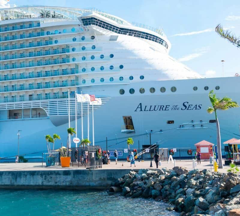 Passengers walk near the massive cruise ship Allure of the Seas docked at a sunny Caribbean port, with palm trees, clear skies, and flags waving near the pier.
