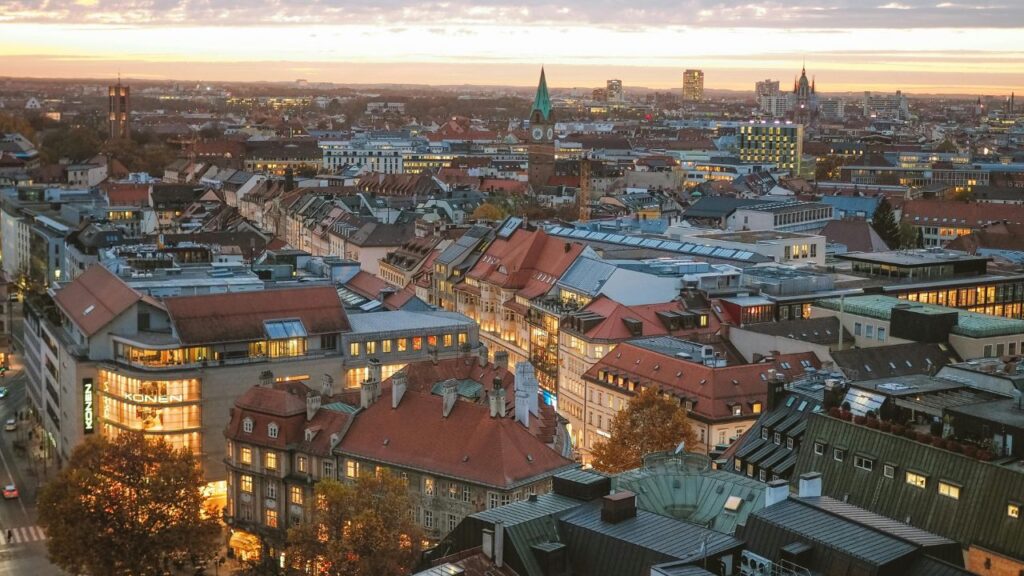 Aerial view of Munich at sunset showing red rooftops, modern buildings, and church spires across the skyline.
