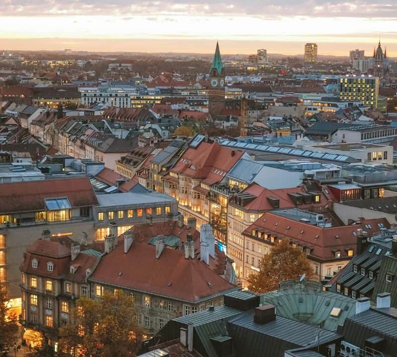 Aerial view of Munich at sunset showing red rooftops, modern buildings, and church spires across the skyline.