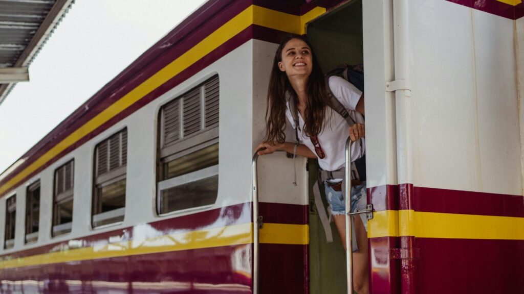 A smiling woman with a backpack leaning out of a train doorway, enjoying the view while standing on the steps of a maroon and yellow train at a station.