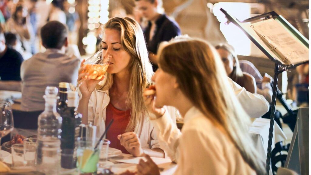 Two women enjoying food and drinks at a lively, crowded restaurant, capturing the experience of discovering a great local dining spot while traveling.
