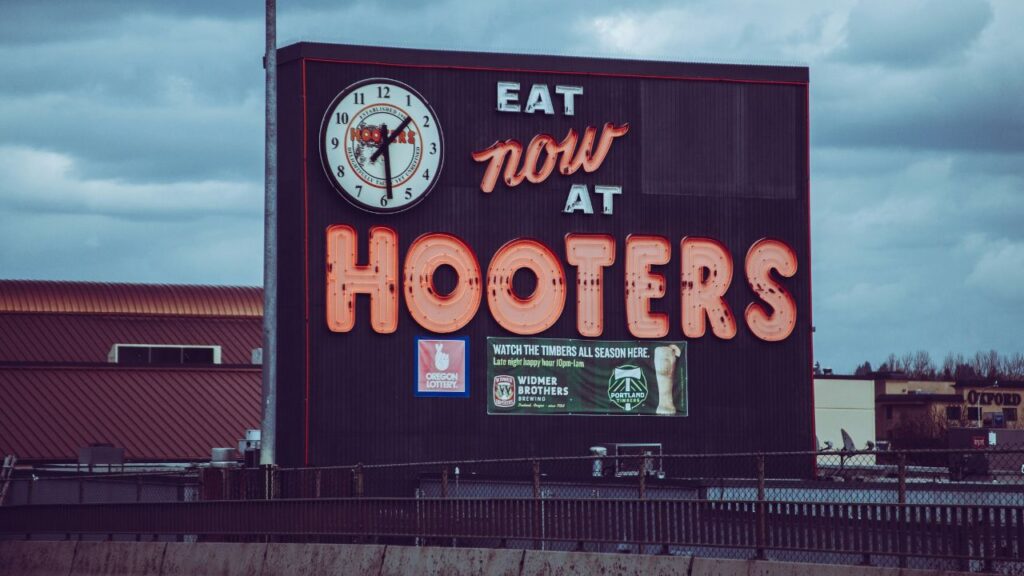A large outdoor sign for Hooters restaurant featuring a clock and neon lettering that reads "Eat Now at Hooters" with logos for Oregon Lottery, Widmer Brothers Brewing, and Portland Timbers.