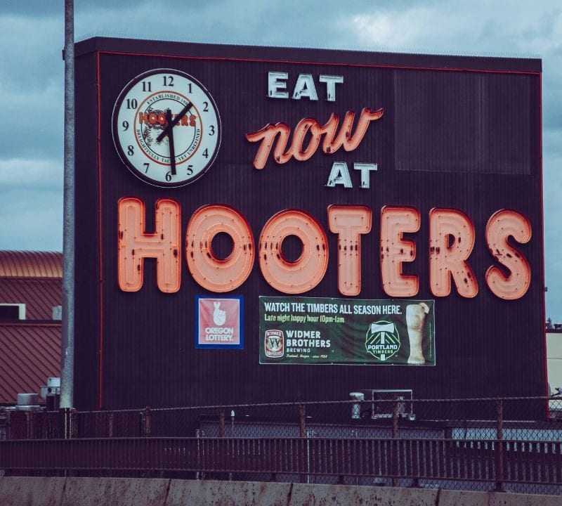 A large outdoor sign for Hooters restaurant featuring a clock and neon lettering that reads "Eat Now at Hooters" with logos for Oregon Lottery, Widmer Brothers Brewing, and Portland Timbers.