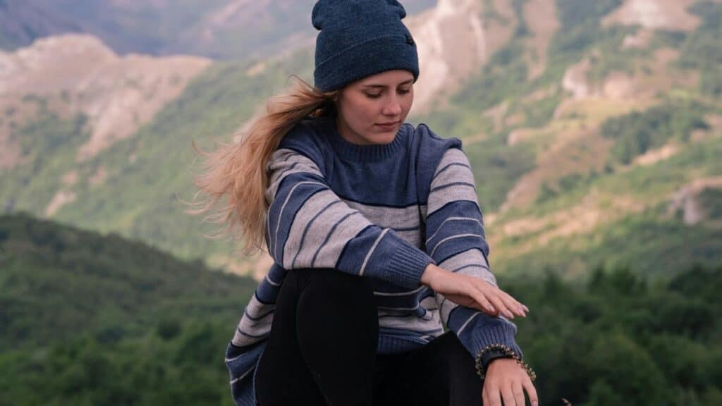 A young woman in a striped sweater and beanie sits alone on a mountain, looking down thoughtfully with wind in her hair.