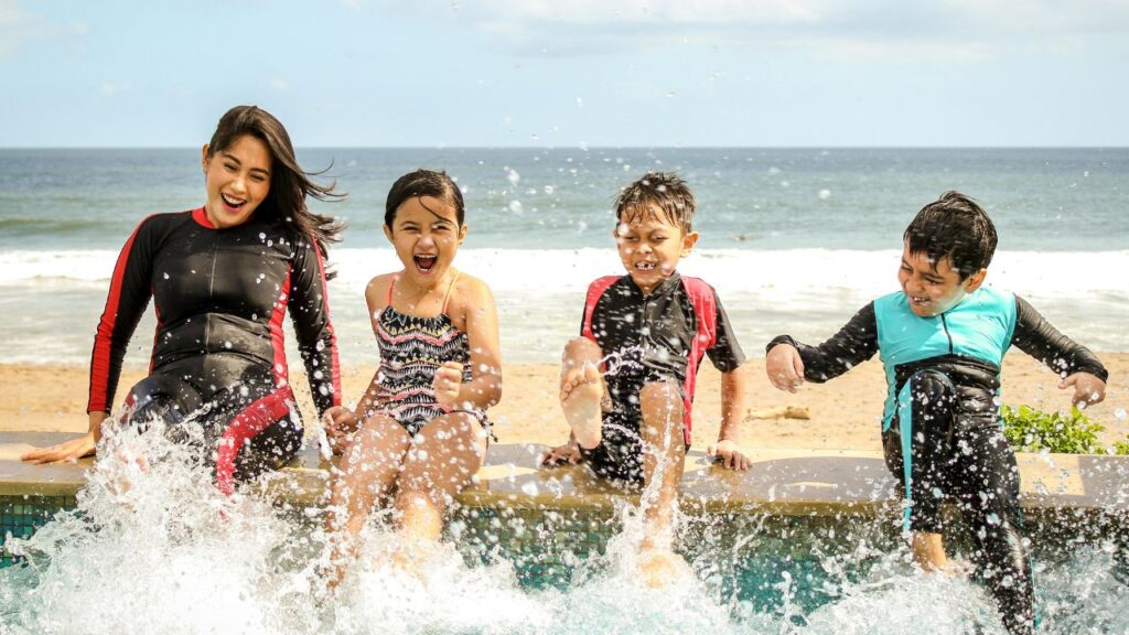 A happy family splashing water by the beach, enjoying a sunny day in swimwear and wetsuits near the ocean.