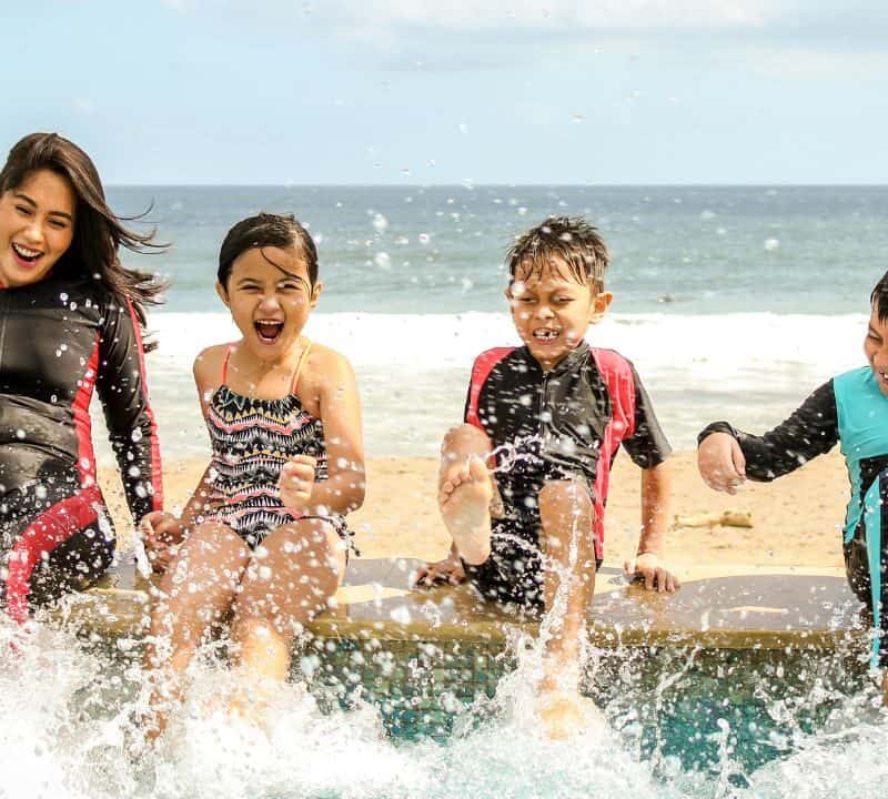 A happy family splashing water by the beach, enjoying a sunny day in swimwear and wetsuits near the ocean.