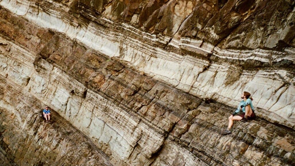 Two people sitting on narrow rock ledges of a steep, layered cliff face, surrounded by dramatic geological formations with horizontal striations.