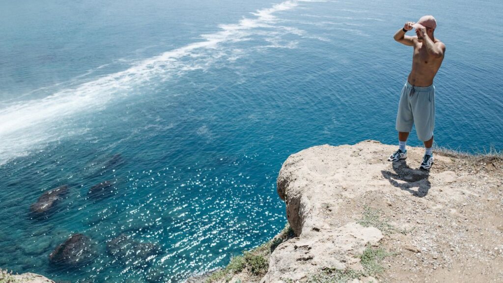 A shirtless man stands alone on a rocky cliff edge, gazing over clear blue ocean water with rocks visible below.