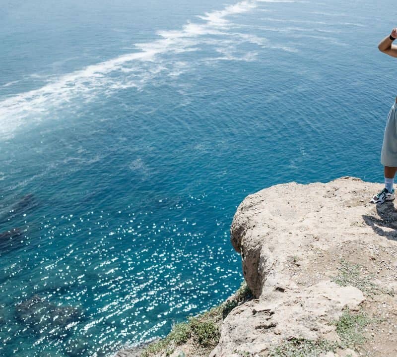 A shirtless man stands alone on a rocky cliff edge, gazing over clear blue ocean water with rocks visible below.