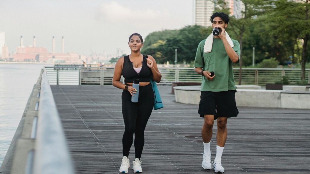 A man and woman in athletic wear walking along a wooden boardwalk by the water, carrying water bottles and a yoga mat, with city buildings in the background.