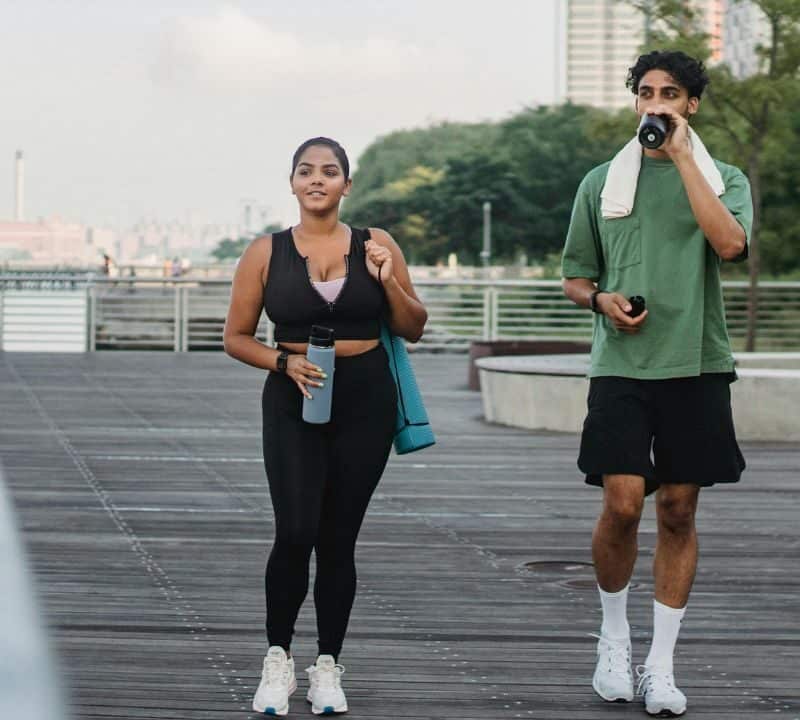 A man and woman in athletic wear walking along a wooden boardwalk by the water, carrying water bottles and a yoga mat, with city buildings in the background.