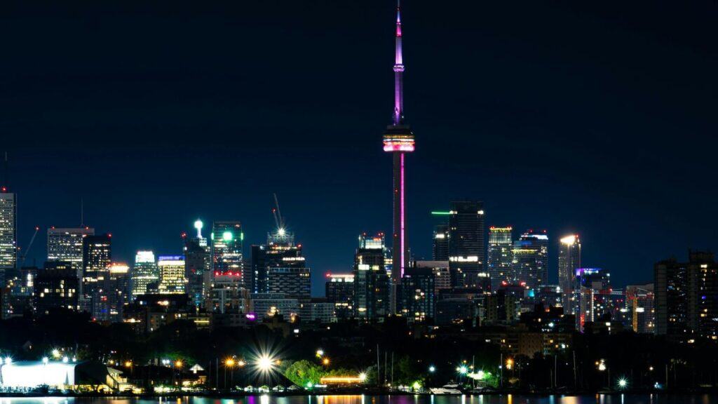 Toronto skyline at night featuring the CN Tower illuminated in pink and surrounded by glowing high-rise buildings.