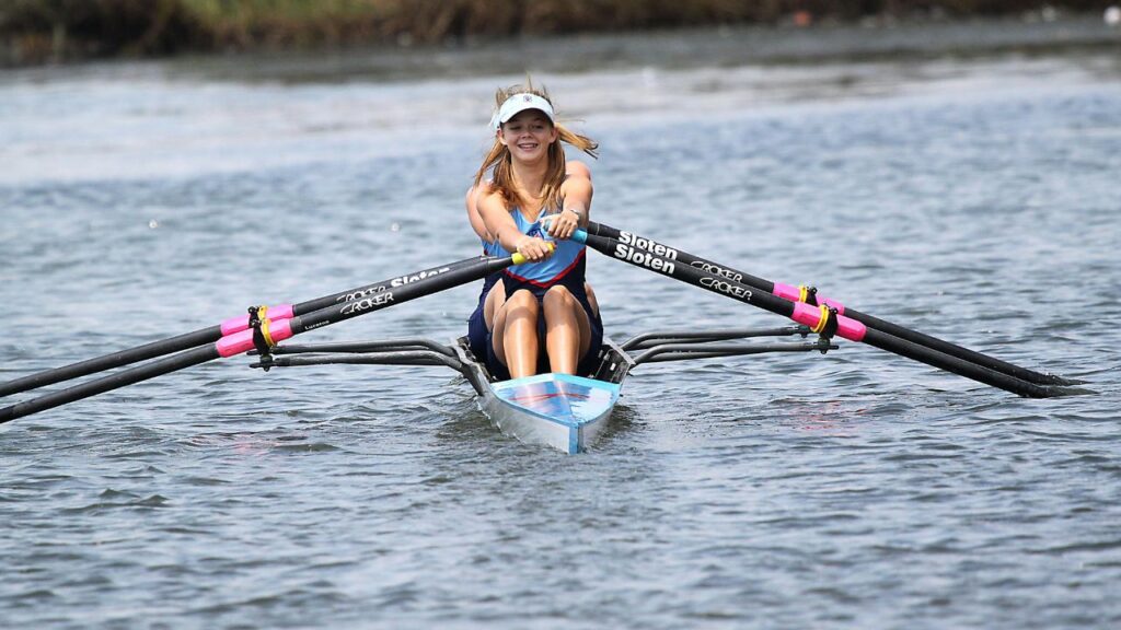 A young woman rows a sculling boat on calm water, smiling as she powers forward with pink-handled oars under a clear sky.