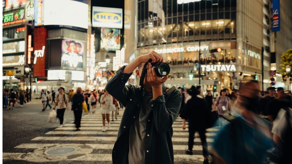 Photographer taking a picture in the middle of Shibuya Crossing at night, surrounded by moving crowds and neon lights.