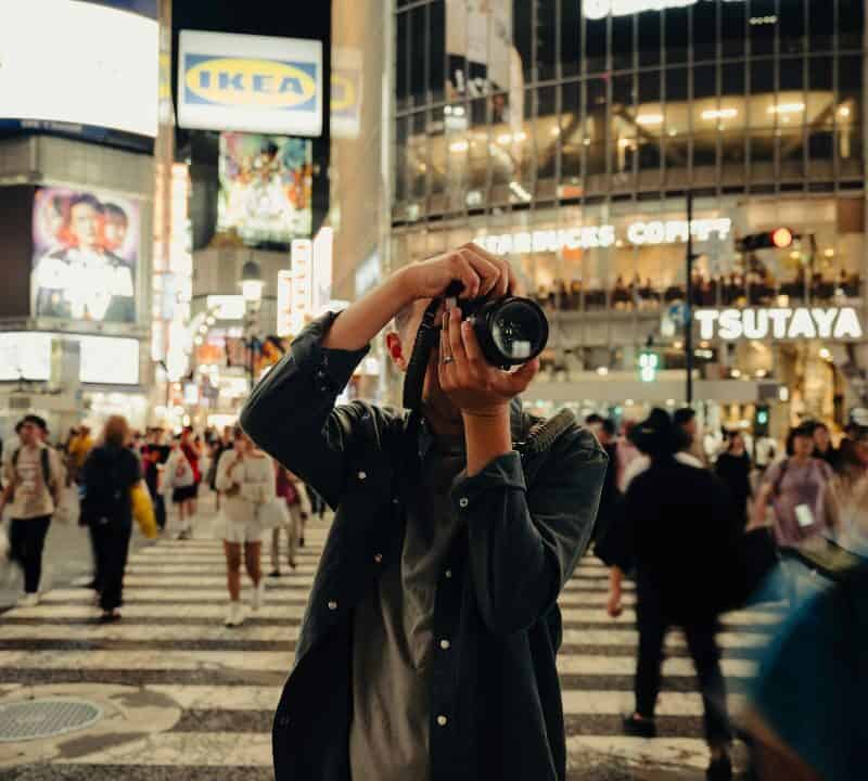 Photographer taking a picture in the middle of Shibuya Crossing at night, surrounded by moving crowds and neon lights.