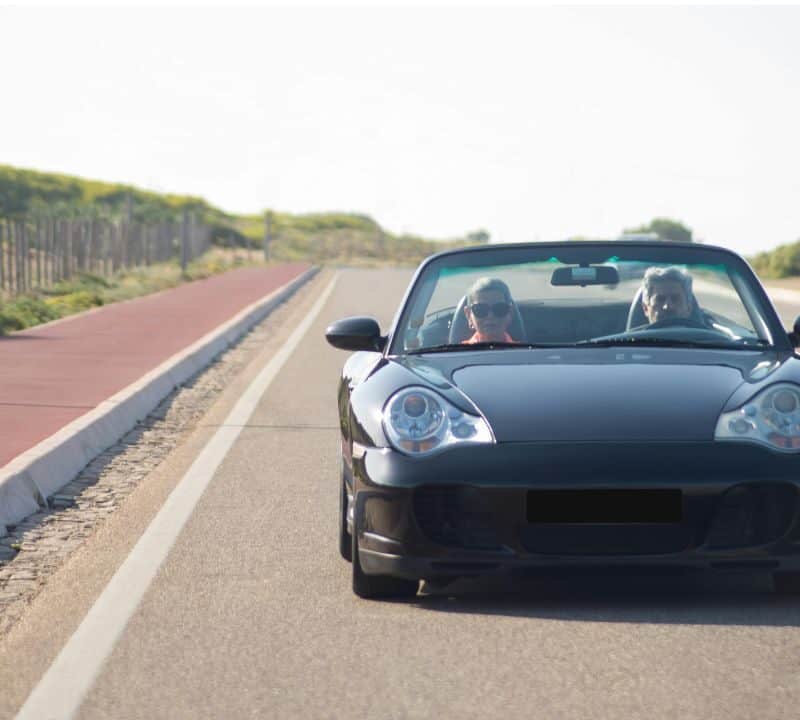 Two people driving a black convertible down an empty scenic road, enjoying a sunny road trip adventure.