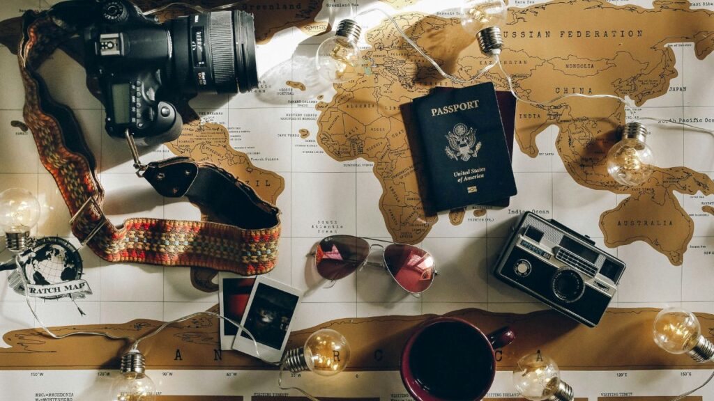 Flat lay of travel items on a world map including a camera, passport, vintage photos, sunglasses, and lightbulbs.