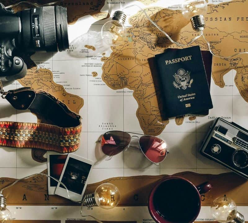 Flat lay of travel items on a world map including a camera, passport, vintage photos, sunglasses, and lightbulbs.