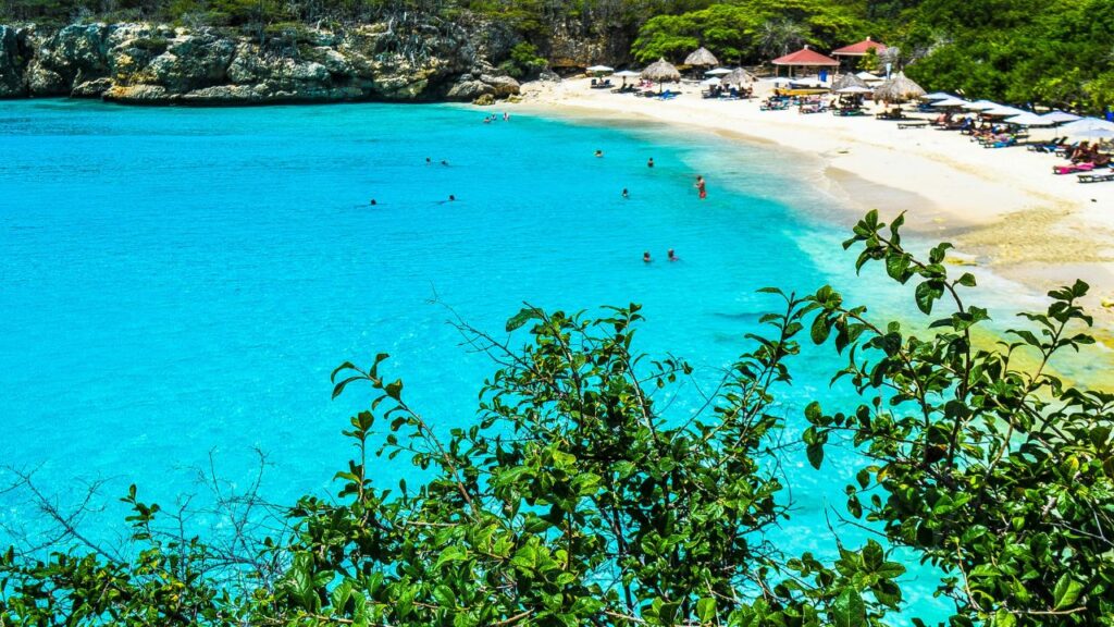 A hidden Caribbean beach with crystal-clear turquoise water, swimmers in the bay, and a quiet white-sand shoreline lined with thatched umbrellas and sunbeds, surrounded by lush green foliage and rocky cliffs.